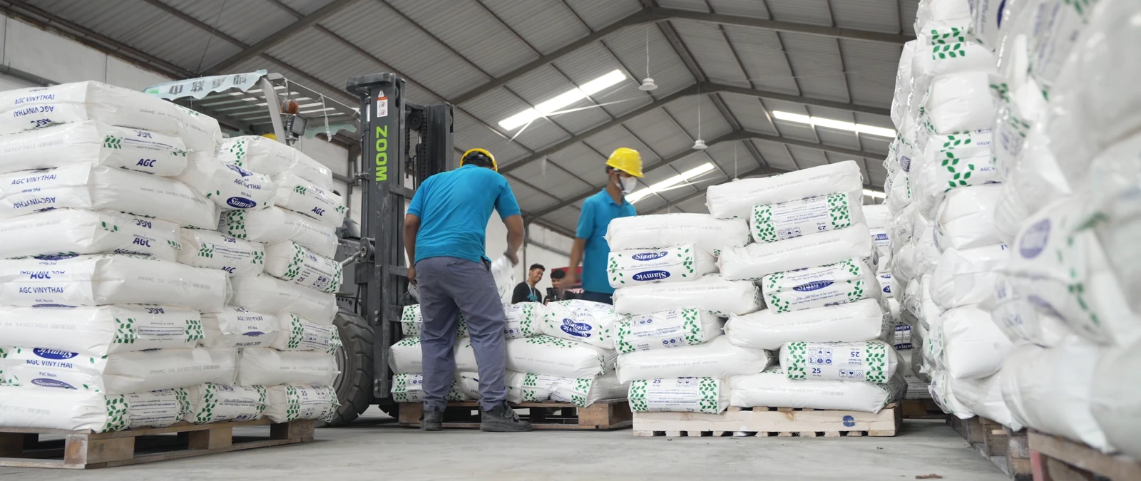 Dua pekerja memindahkan karung bahan di antara tumpukan palet gudang.